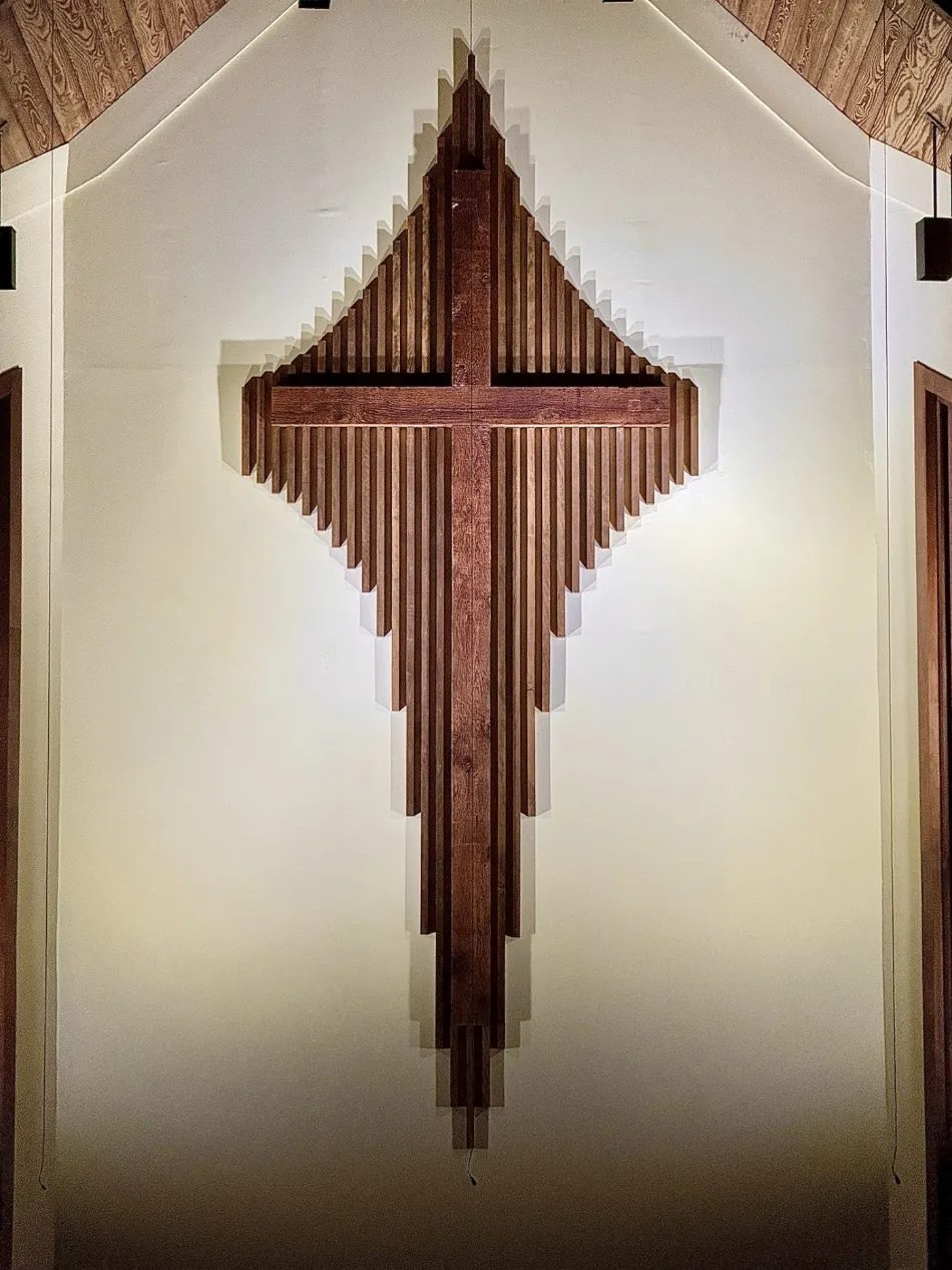 The wooden chancel cross of the sanctuary — a sunburst of graduated slats around the cross — lit warmly against the wall beneath the vaulted ceiling