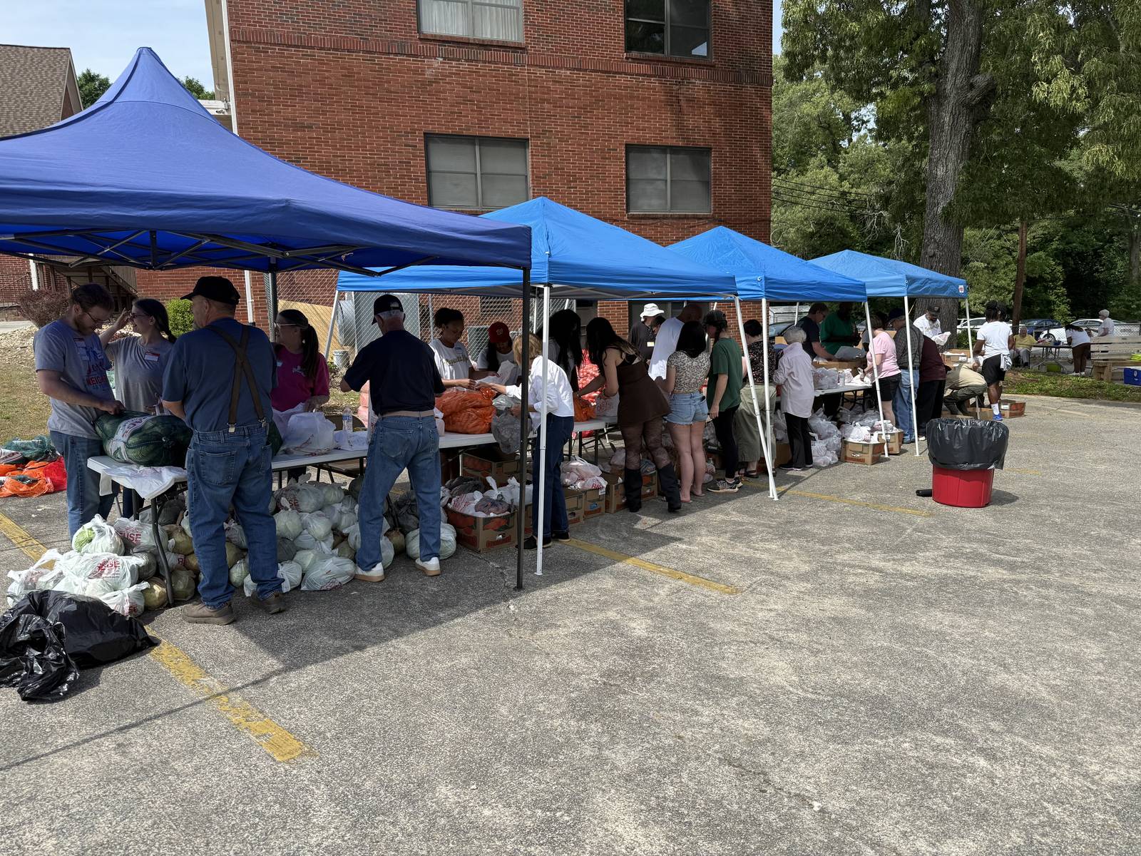 Volunteers working under a line of blue canopies at a drive-thru food pantry distribution in the church parking lot, pallets of produce beside them and the three-story education building behind